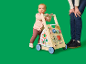 Baby spielt mit einem hölzernen Aktivitäts-Lauflernwagen, ein Erwachsener steht daneben.