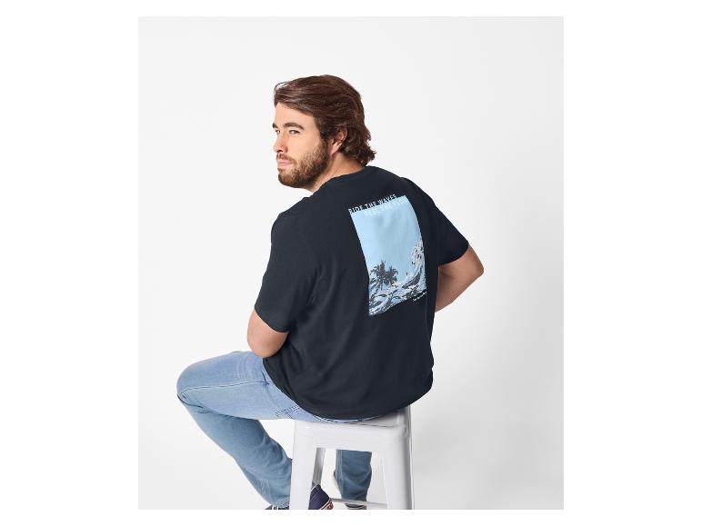 Mann in schwarzem T-Shirt mit Strandaufdruck und Jeans, auf einem Hocker sitzend.