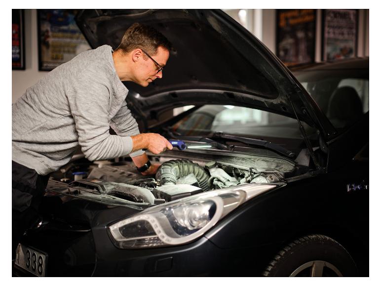 Mann mit Arbeitsleuchte »The Mechanic« inspiziert Automotor.