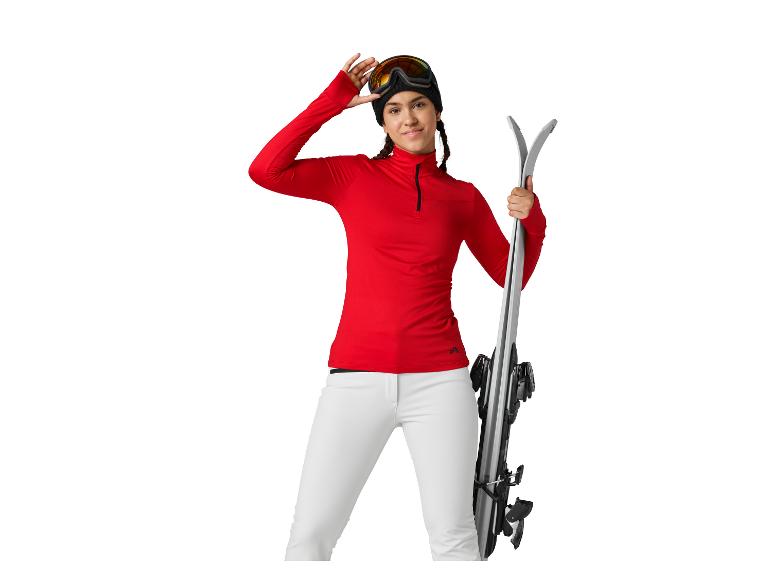 Frau in rotem Skishirt und weißer Hose hält Skier und Skibrille.