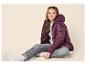 Lächelndes Mädchen in einer pflaumenfarbenen Daunenjacke, grauem Pullover und Jeans, auf dem Boden sitzend.