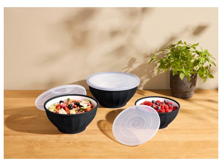 Drei schwarze Schüsseln mit transparenten Deckeln, gefüllt mit Salat und Beeren, auf einem Holztisch.