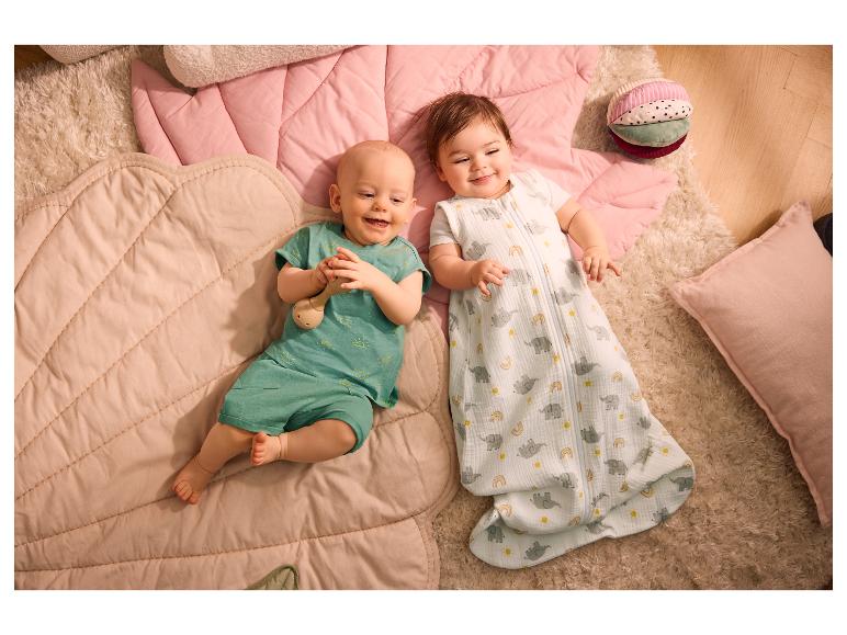Zwei lächelnde Babys liegen auf rosa Spielmatten, eines im Schlafsack, das andere in einem grünen Outfit.