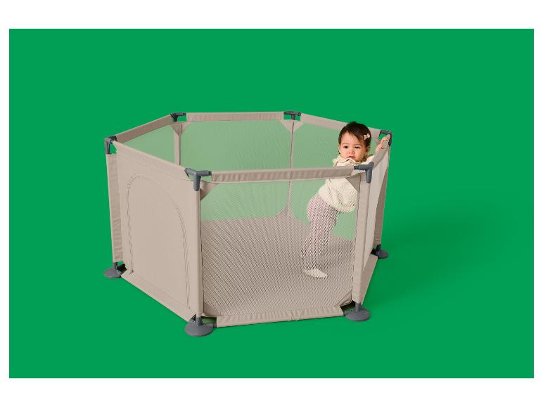 Baby in einem sechseckigen, beigen Laufstall mit Netzwänden, stehend und am Rand lehnend.