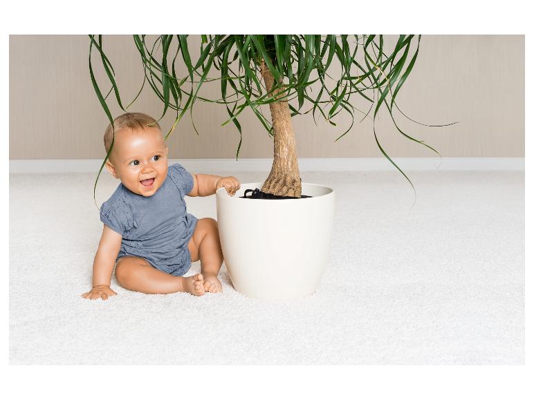 Ein lächelndes Baby in Blau sitzt neben einem Pflanzentopf.