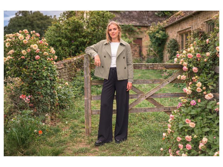 Frau in grüner Jacke und schwarzer weiter Hose an einem Holztor mit Rosen.