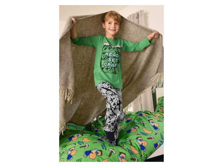 Junge in grünem Minecraft-Pyjama mit „READ, SET, CRAFT“-Aufschrift, stehend auf einer Minecraft-Bettdecke.