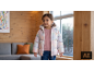 Mädchen in rosa Pullover und Pufferjacke, stehend in einem Raum mit Holzwänden.