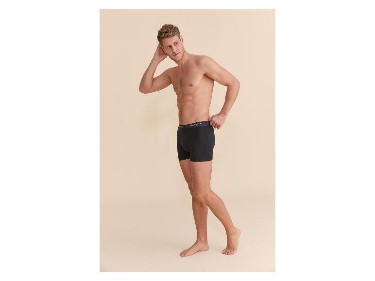 Blonder Mann in schwarzen Boxershorts vor beigem Hintergrund.