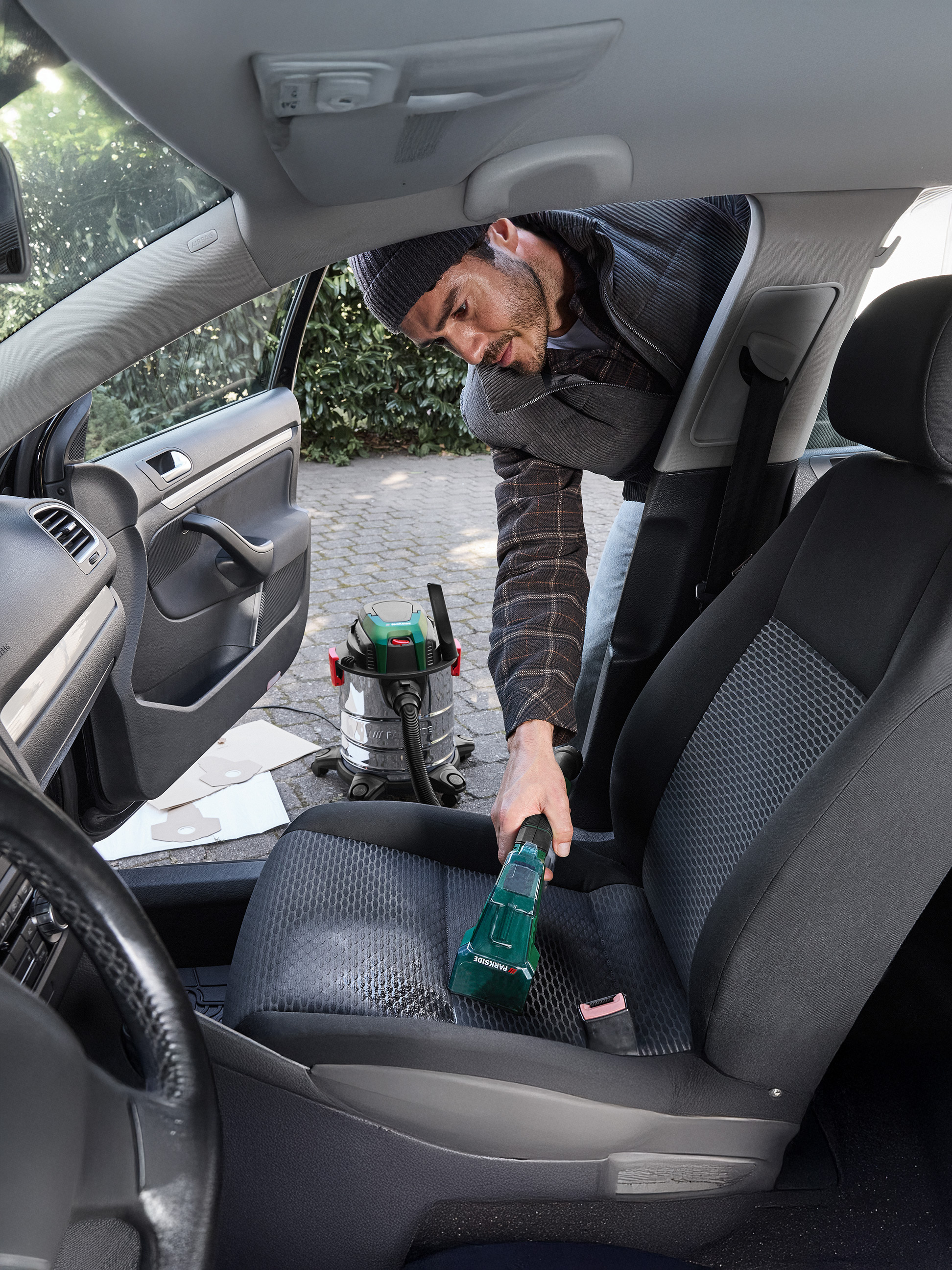 Mann reinigt Autositz mit einem Parkside Handstaubsauger, mit einem Nass-/Trockensauger im Hintergrund.