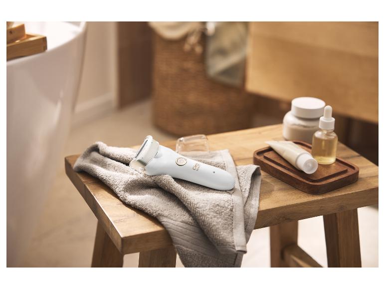 Damen-Epilierer auf einem Handtuch, daneben Hautpflegeprodukte auf einem Holzhocker.