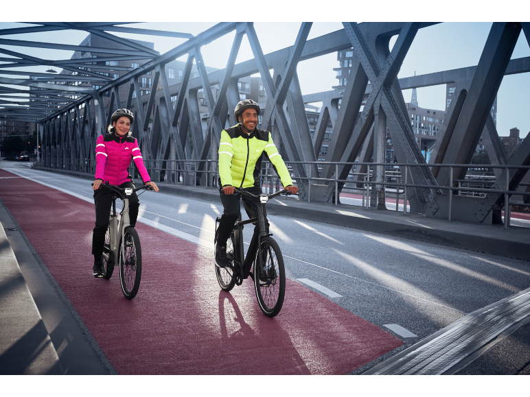 Zwei Personen in reflektierenden Fahrradjacken und Helmen fahren E-Bikes auf einer Brücke.