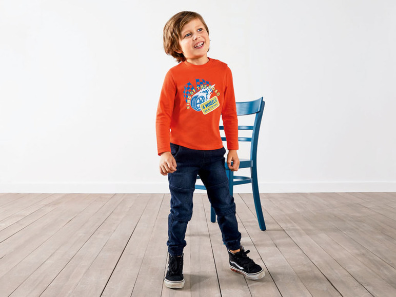 Junge in einem orangefarbenen T-Shirt mit Rennauto-Aufdruck, blauen Jeans und schwarzen Sneakern.