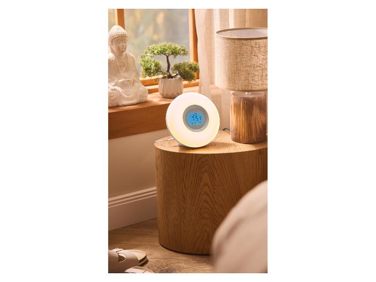 Lichtwecker auf Nachttisch mit Lampe und Dekorationen, Fenster im Hintergrund.