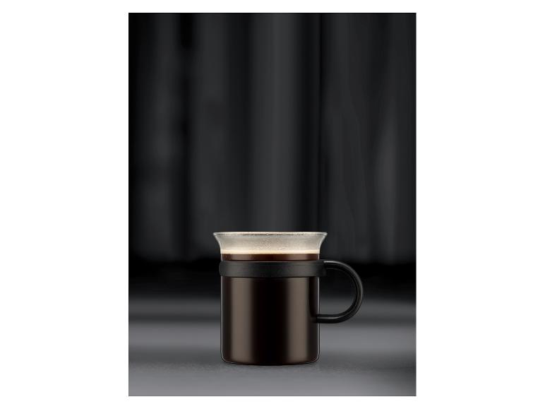 Kaffeebecher aus Glas und Metall mit schwarzem Griff auf dunklem Hintergrund.