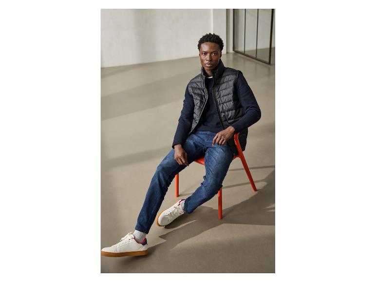 Mann in schwarzer Weste, marineblauem Pullover und Jeans, sitzt auf einem orangefarbenen Stuhl.