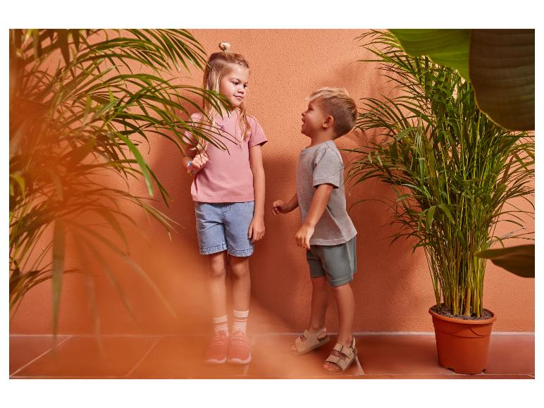 Zwei Kinder in lässiger Sommerkleidung, ein Mädchen in einem rosa T-Shirt und Jeansshorts, ein Junge in einem grauen T-Shirt und grünen Shorts.