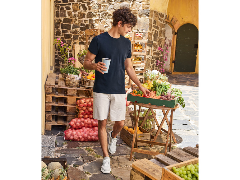 Mann mit einer Thermoskanne und weißen Shorts geht an Obst- und Gemüseständen vorbei.