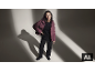 Eine Frau in einer malvenfarbenen Daunenjacke, einem schwarzen Pullover und einer schwarzen Hose posiert in einem Studio.