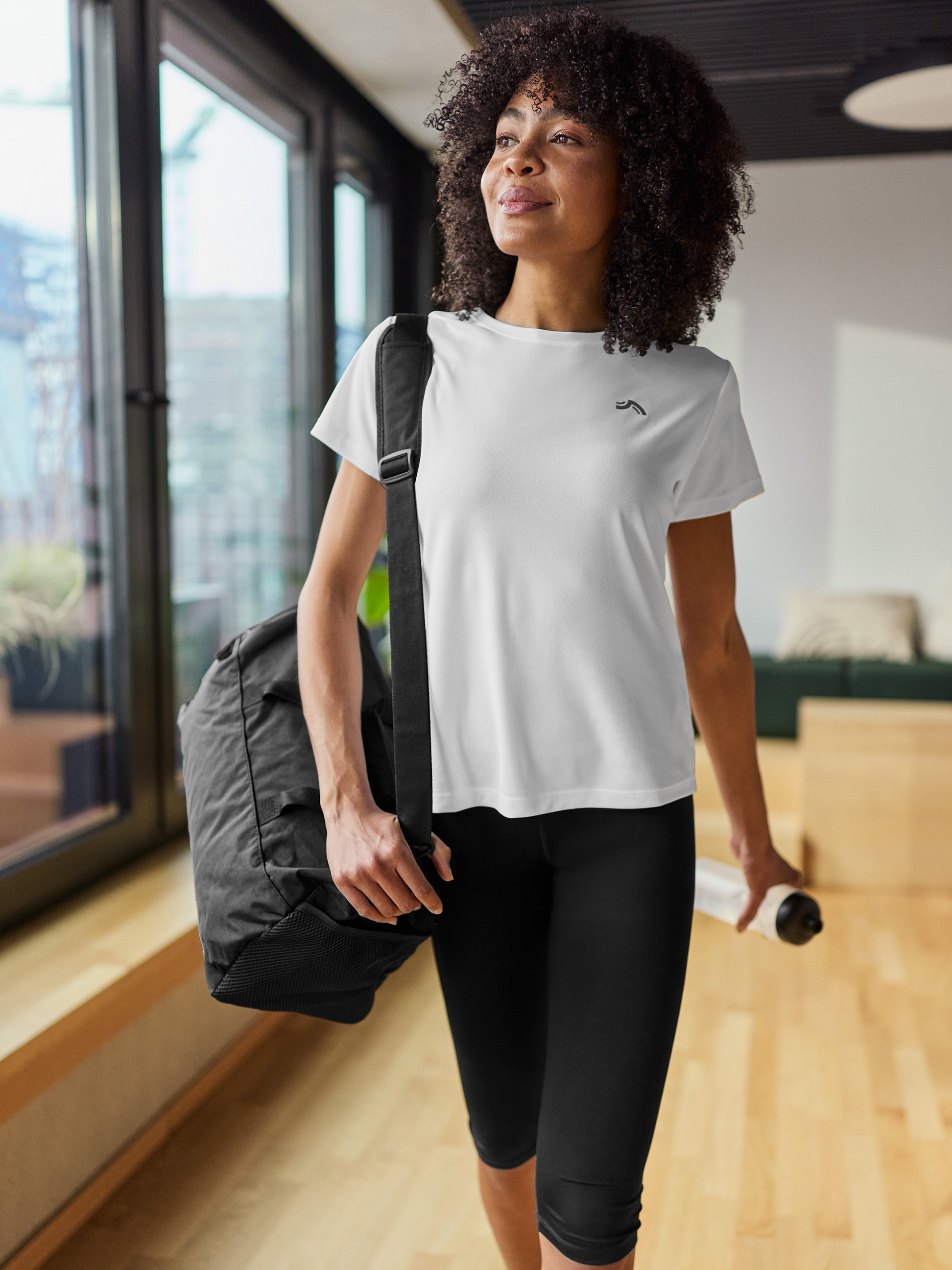 Frau in weißem Sport-T-Shirt, schwarzen Leggings, mit Sporttasche und Wasserflasche.