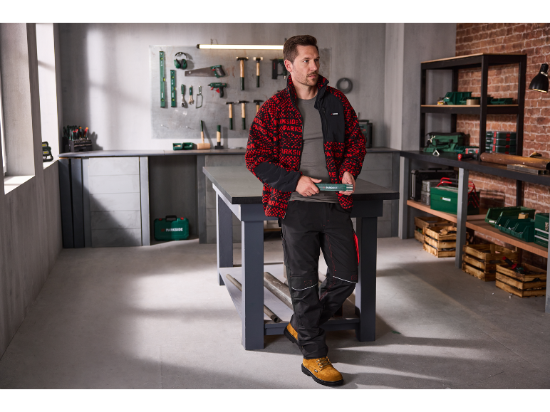 Mann in rot-schwarzer Fleecejacke und Arbeitshose in einer Werkstatt.