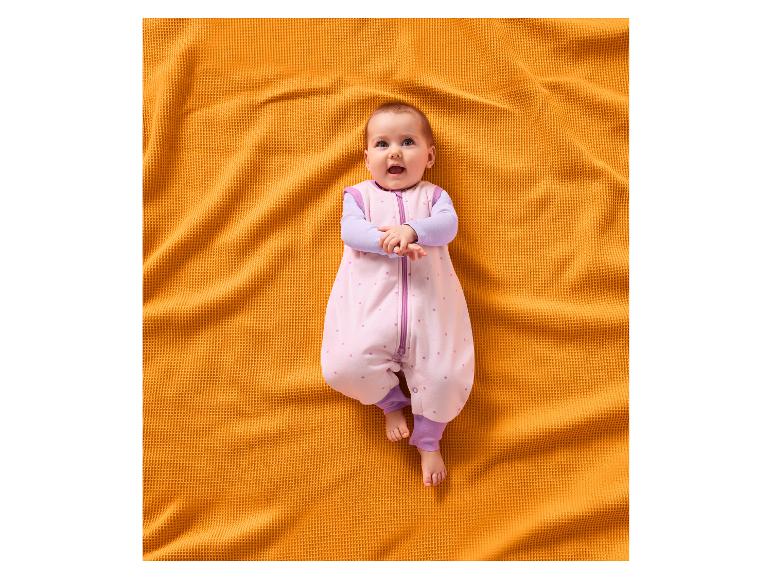 Lächelndes Baby in einem rosa Schlafsack mit Sternen, liegend auf einer orangefarbenen Decke.