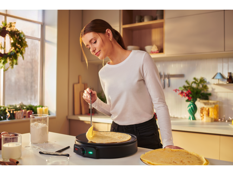 Frau macht Crêpes auf einem Crêpes-Maker in der Küche, mit Zutaten und Weihnachtsdeko.