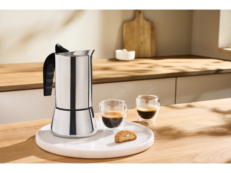 Edelstahl-Espressokocher mit zwei Espressotassen und einem Biscotti auf einem Marmortablett.