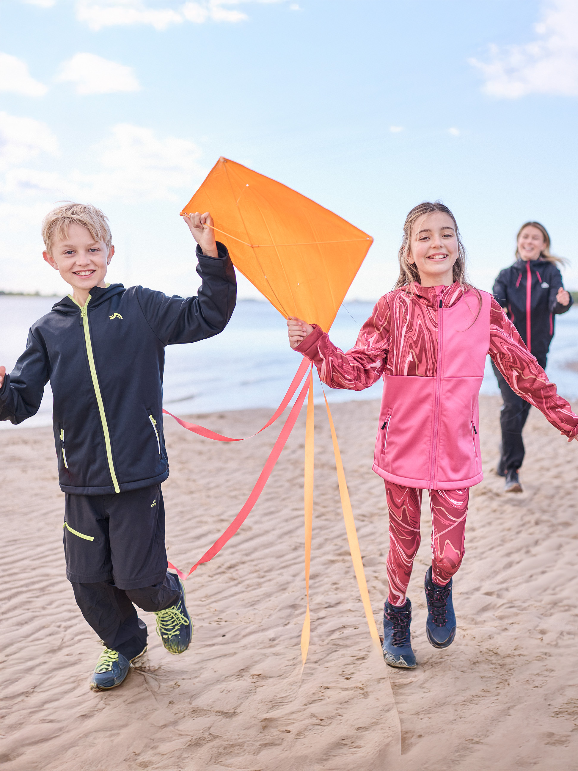 Kinder in Sportkleidung rennen am Strand mit einem Drachen, Frau im Hintergrund.