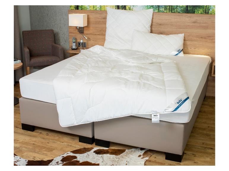Ein Bett mit weißer BeCo-Bettwäsche, einem Nachttisch und einem Sessel in einem Zimmer.