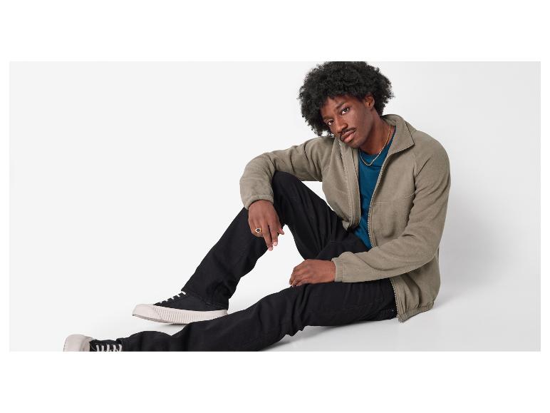 Mann in einer khakifarbenen Fleecejacke, blauem T-Shirt und schwarzen Jeans.