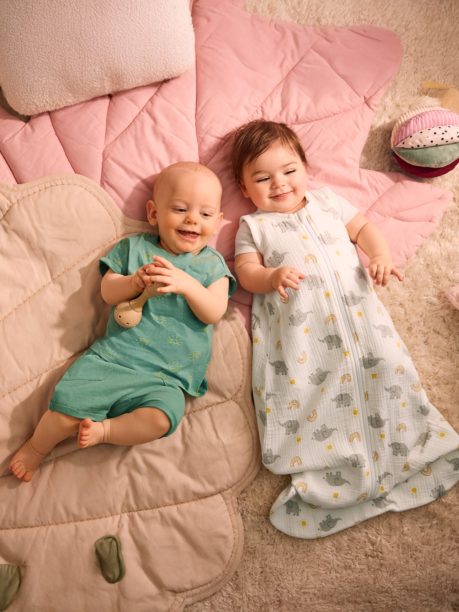 Zwei Babys liegen auf einer rosa Decke, eines in grüner Kleidung, das andere in einem Schlafsack mit Elefanten.