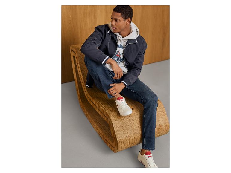 Mann sitzt in einer Sweat-Denim-Jeans, blauer Jacke und weißen Sneakern.