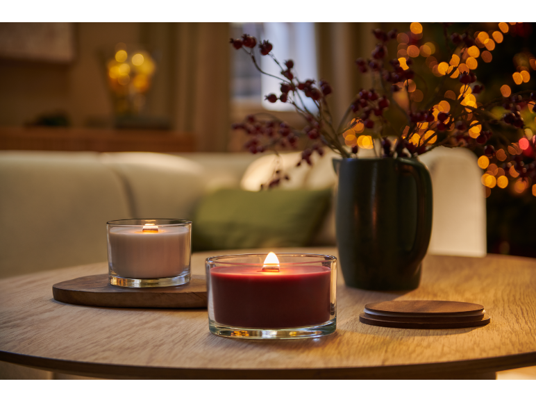 Zwei brennende Kerzen auf einem Holztisch, mit einer Vase voller Beeren und verschwommenen Lichtern im Hintergrund.