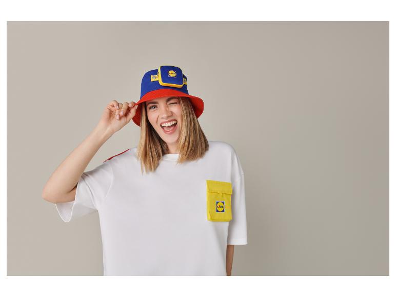Junge Frau mit blau-rotem Fischerhut, weißem T-Shirt mit gelber Tasche, zwinkernd und lächelnd.