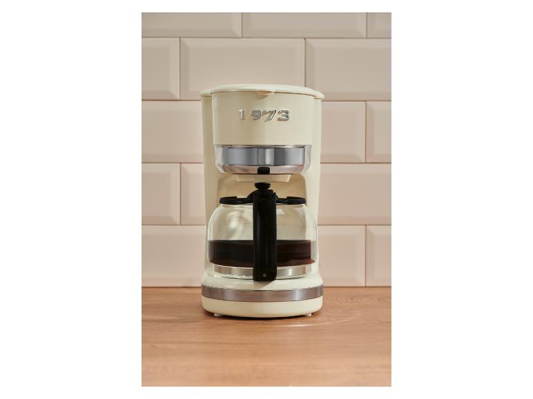 Cremefarbene Kaffeemaschine mit Kanne auf einer Holztheke, vor beigen Fliesen.
