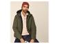 Mann in grüner Steppjacke, beigem Half-Zip-Pullover, dunklen Jeans und roter Mütze.