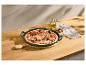 Pizza mit Schinken, Oliven und Pilzen in einer grünen Backform auf einem Holztisch.