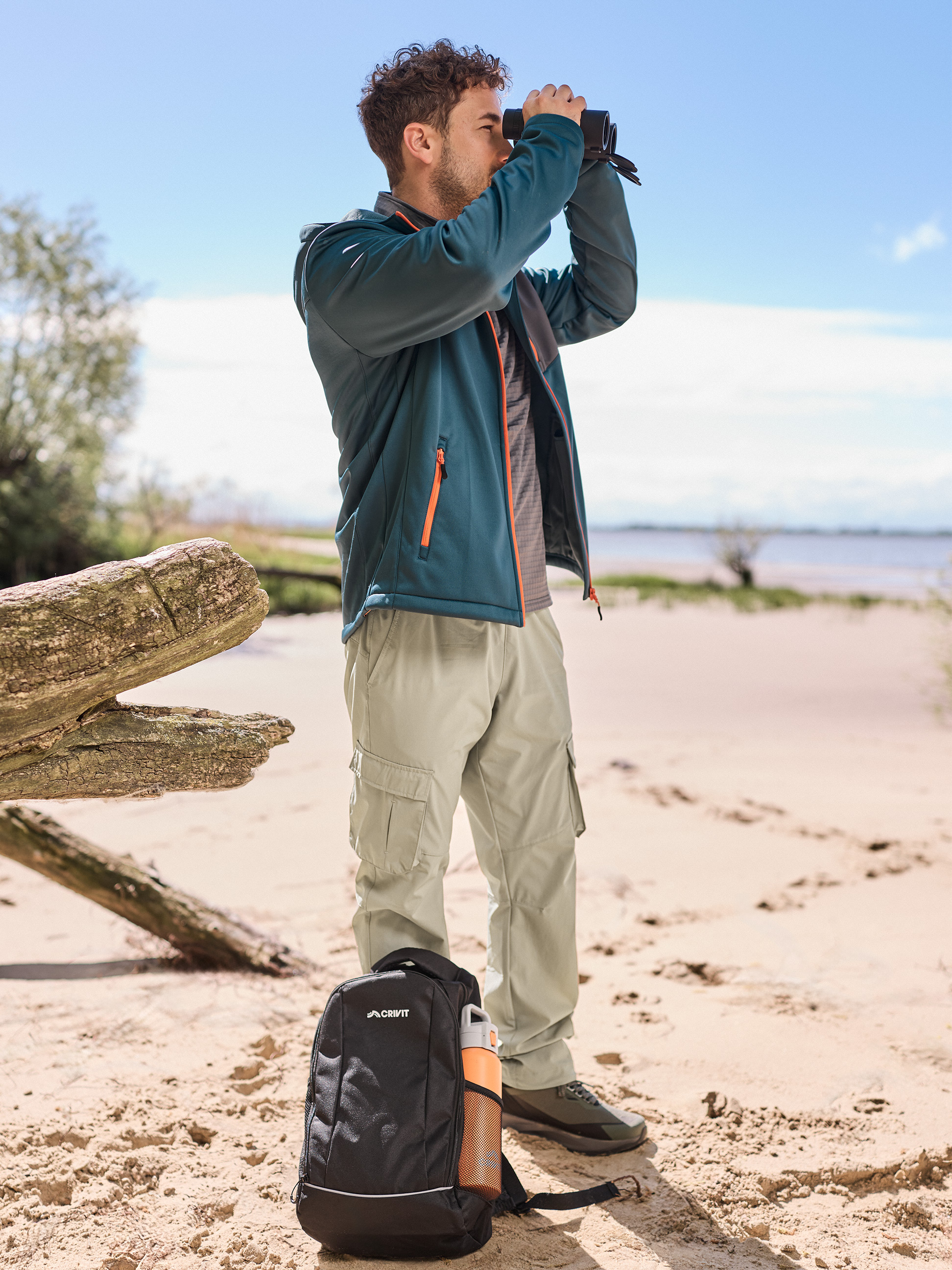 Mann in Wanderjacke und -hose, Fernglas, Rucksack und Wasserflasche am Strand