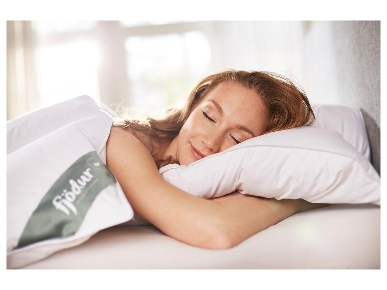 Frau schläft auf weißem Kissen und Decke mit Fjödur-Logo in sonnigem Schlafzimmer.