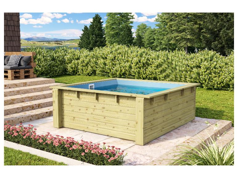 Quadratischer Holzpool mit blauem Wasser in einem Garten mit Büschen und Palettensofa.