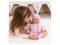 Mädchen spielt mit einer Babypuppe in rosa Kleidung auf einem weißen Teppich.