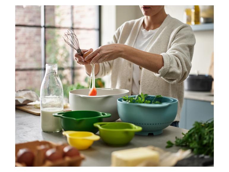 Eine Person schlägt ein Ei in eine Schüssel mit Küchenutensilien und Zutaten auf dem Tisch.