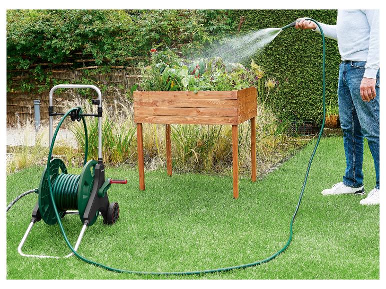 Mann bewässert Pflanzen in einem Hochbeet mit einem grünen Gartenschlauch auf einem Wagen.