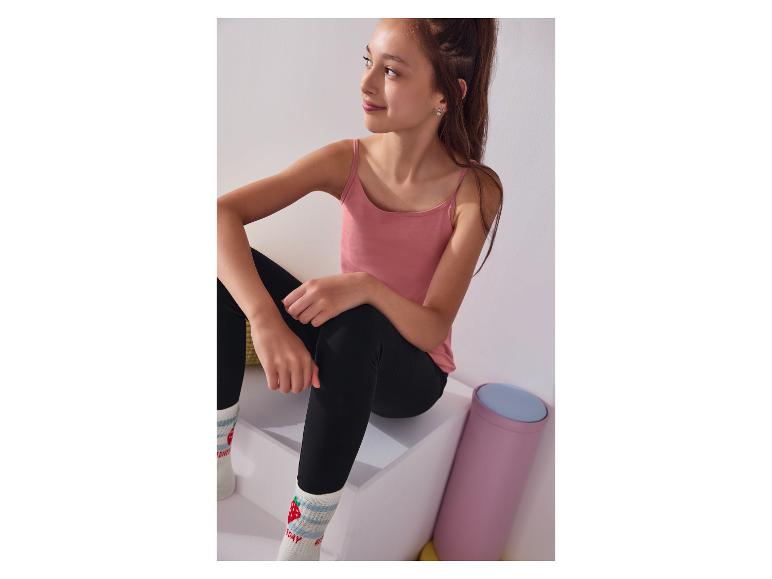 Ein Mädchen lächelt, trägt ein rosa Top, schwarze Leggings und Erdbeersocken.