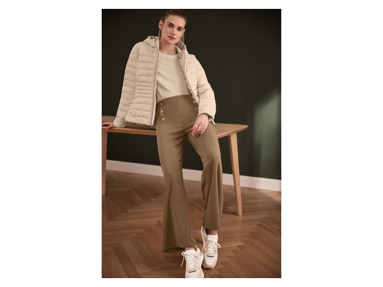 Frau in beiger Jacke, cremefarbenem Shirt und brauner Hose mit goldenen Knöpfen.