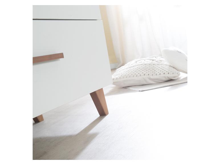 Weiße Kommode mit Holzgriffen und -beinen, daneben ein gestricktes Kissen auf dem Boden.