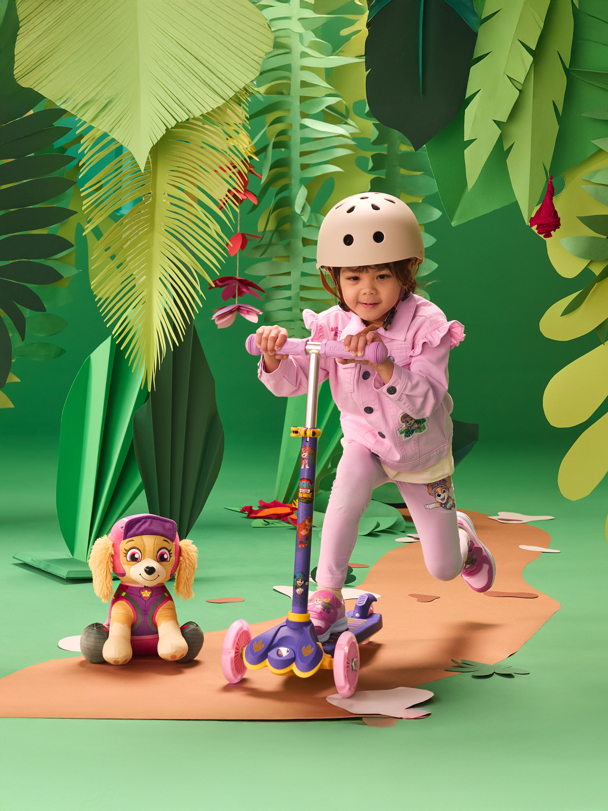 Mädchen mit Helm auf Roller, mit Plüschtier, in Dschungel-Kulisse.