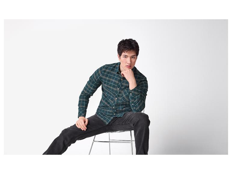 Mann in grünem Karohemd und schwarzer Jeans, auf einem Hocker sitzend.