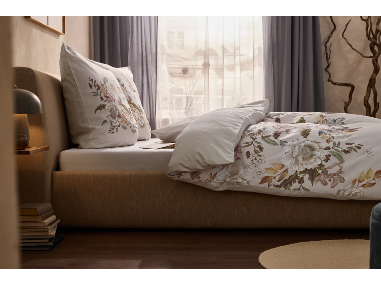 Schlafzimmer mit floraler Bettwäsche und Kissen, Nachttischlampe und Büchern.
